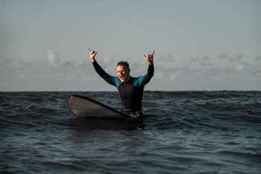 Dalgıç kıyafetli, gülümseyen genç sörfçü. Sörf tahtasının üzerinde oturuyor. Elleri ve ağzı açık.