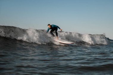 Dalgıç kıyafetli yakışıklı genç sörfçü sörf tahtasının üzerinde ona doğru yaklaşan bir dalgayla ayağa kalkar.