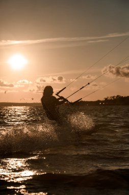 Dişi wakeboardcu, gün batımına karşı üç kabloyu sıkıca tutarak denizde bir wakeboard sürüyor.