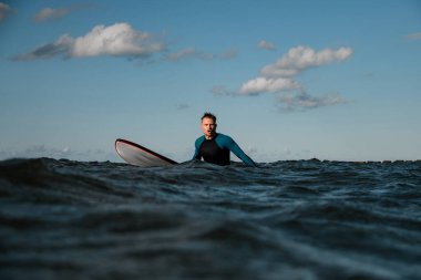 Genç erkek sörfçü denizde sörf tahtasını itiyor ve kameraya bakarken bağırıyor.
