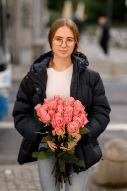 Güzel bir buket pembe gül. Gri şehir caddesinde kapüşonlu ve gözlüklü siyah ceketli bir kız.