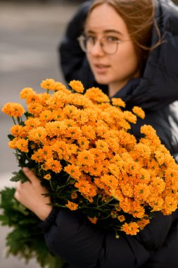 Gözlüklü ve kapüşonlu siyah ceketli bir kız, sarı çiçekli bir buket şehir caddesinde duruyor, koyu renk saçlı güzel bir kızın fotoğrafı.