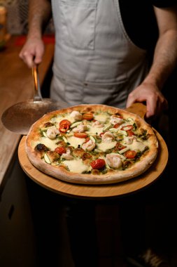 Aşçı gururla soyulmuş karidesle süslenmiş bitmiş pizzayı sunar. Kırsal ahşap bir tahtada nefis bir mutfak zevki için sunar..