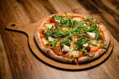 Bu yakın çekim bitirilmiş bir pizzanın zarif sunumunu yakalar, roka ile ustalıkla süslenir ve kırsal ahşap bir tahtada sergilenir, hem görsel hem de yemek pişirme zevki sunar..