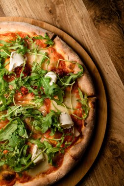 Bu yakın plan roka ile süslenmiş bitmiş bir pizzanın detaylı bir perspektifini sunuyor. Kırsal ahşap bir tahtanın üzerinde zarif bir şekilde sergilenerek bir mutfak başyapıtı yaratıyor..