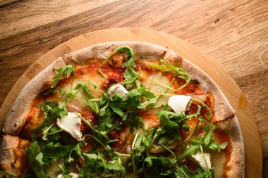 Bu detaylı yakın planda, bitirilmiş pizza cömert bir roka yardımıyla geliştiriliyor, kırsal ahşap bir tahtada özenle sunuluyor, aşçılık mükemmelliği elde ediliyor..