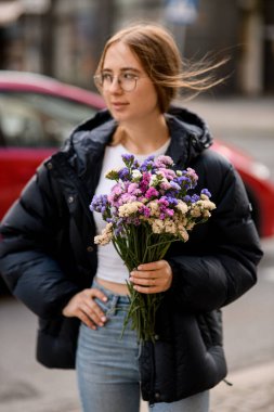 Gözlüklü güzel bir kızın ellerinde mor-pembe-beyaz-bej çiçekler var. Gün boyunca siyah ceketli bir caddede duruyor.