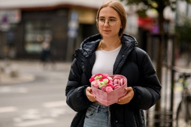 Gözlüklü kızın elinde kalp şeklinde pembe bir demet badem ve çiçekler ve şehrin ön tarafında duran siyah ceketli kız.