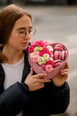 Gözlüklü, siyah saçlı bir kız şehir caddesinde duruyor, gri bir arka planda, kalp şeklinde bir buketle.