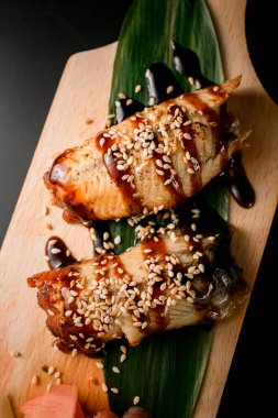 Büyük yeşil bir yaprağın üzerinde koyu kahverengi soya soslu balık ve siyah arka planda ahşap bir tahta.