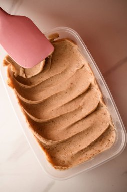 Plastik bir kapta, açık kahverengi krem, bej masa arkasındaki silikon bir spatula yardımıyla alta dalgalı bir şekilde yayılır.