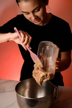 Kadın pastacı, plastik bir kabın içinden durağan bir mikserin metal kasesine açık kahverengi krema döker.