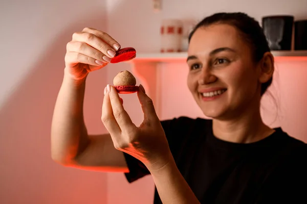 Smiling woman pastry chef holding two halves of red macaroon in front of her face with beige and red cream to seal them