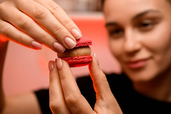 Smiling woman confectioner with French manicure holds two halves of red macaroon with beige cream, holding them in front of her face