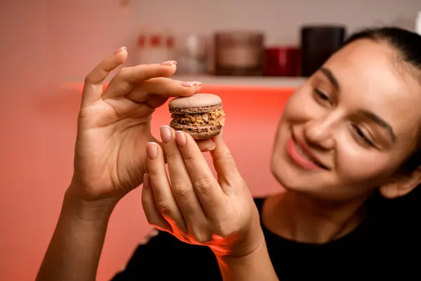 Smiling female pastry chef is admiring a brown macaron decorated with cornflakes that she has made and is holding in her hand