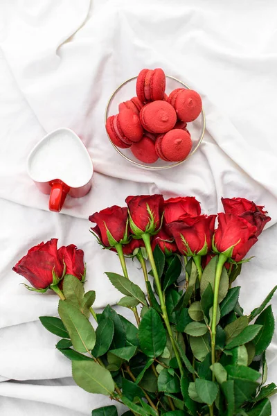 Beautiful bouquet of roses and delicious macaroons in a glass plate on a white fabric background. Tenderness red roses on a white satin