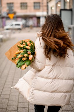 Sırtını kolluyor. Beyaz ceketli, koyu kahverengi saçlı, şefkatli bir kızın ellerinde pembe laleler.