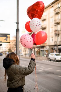 Gri ceketli, kapüşonlu ve siyah pantolonlu bir kızın ellerinde pembe ve beyaz balonlar.