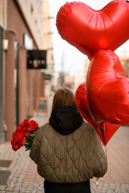 Gri ceketli, siyah pantolonlu, bir sonbahar günü sokakta duran kızın ellerinde kırmızı kalp şeklinde balonlar.