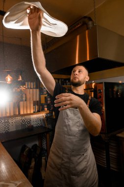 Üniformalı adam pizza hamuru yapıyor. Aşçı mutfakta elinde yuvarlak bir pizza tabanı döndürür.