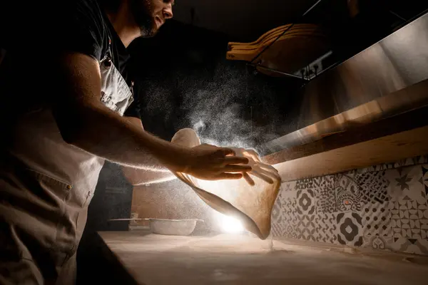 Dark kitchen in white flour and man in gray apron making a pizza dough on a table before preparing a pizza