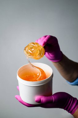 Plastik bir kapta parlak bal şeffaf macunu mavi gömlekli ve arka planda pembe eldivenli bir uzmanın ellerinde.