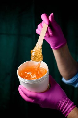 Mavi gömlekli ve pembe eldivenli bir kadının ellerinde tahta bir spatulayla plastik bir kapta parlak şeffaf şeker yapıştırıcısı.