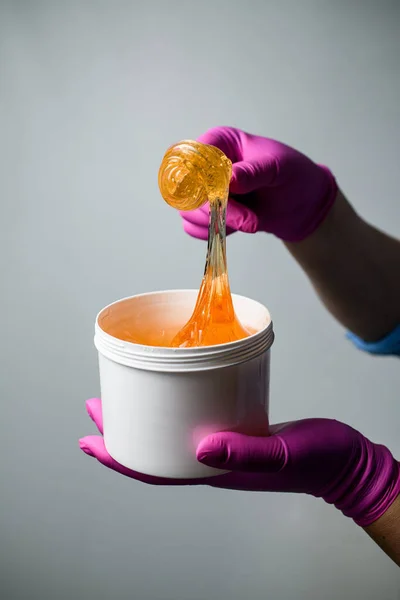 Bright honey-brown sugaring paste in a plastic container with a wooden spatula in hands in blue shirt and pink gloves