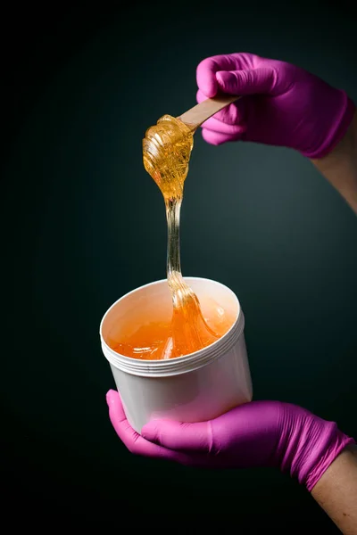 Dark green background and sugaring paste paste for depilation with a wooden spatula in a cosmetologists hands in a pink gloves