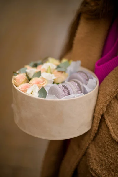 身穿棕色外套的女性手拿着一个装有粉红玫瑰和紫色马卡龙的礼品盒 — 图库照片