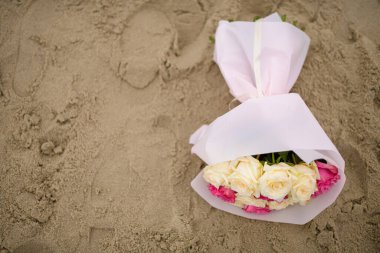Beyaz bir ambalajda beyaz ve pembe gül buketi deniz kıyısındaki ıslak kumun üzerinde yatıyor.