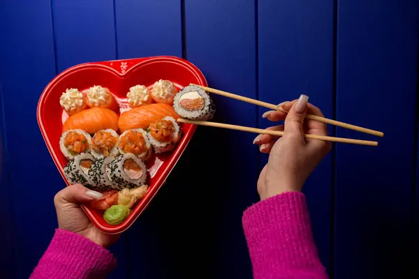 Female hands holding sushi rolls with chopsticks over a heart-shaped sushi box on a blue wall background