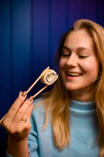 stock image Young pretty smiling woman holding sushi roll with chopsticks and looking at it on blue background