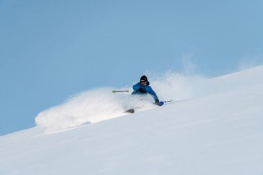 Mavi kayak takımlı kayakçı elinde direklerle yamaçtan aşağı iniyor ve kar yukarı doğru yükseliyor.