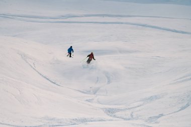 Kayak takımlı iki kayakçı ellerinde sırıkla karlı bir yamaçtan aşağı ortak bir inişi tamamlıyorlar, dikiz manzarası, toz kar yükseliyor.