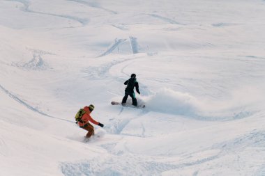 Tam teçhizatlı iki snowboardcu, yamaçtan yokuşa atılıyorlar. Diğer kayakçıların izleri önümüzde görünüyor.