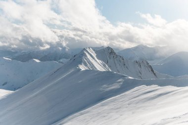 Yüksek dağların karlı zirveleri, beyaz bulutlu, kenar manzaralı mavi gökyüzünün arka planında görülebilir.
