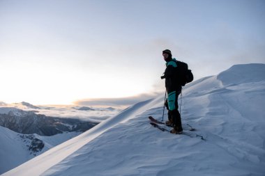 Kayakçı bir dağ yamacına tırmanıyor, sis örtüsü altında kalan alçak dağların arka planına karşı, başka bir kayakçı neredeyse en tepede.