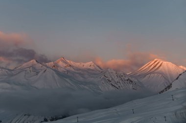 Uzak bir dağ kitlesi manzarası, bazı zirveleri güneşin batışının son ışınlarıyla kırmızı bir gökyüzünün arka planında yıkanmış.