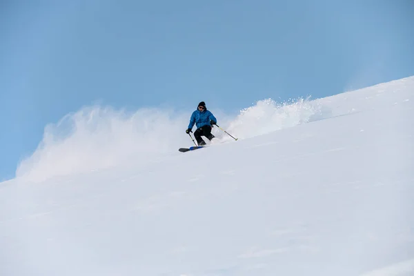 穿着蓝色夹克 手里拿着杆子的男滑雪者从雪坡上滑下去 雪粉就会侧身飞走 — 图库照片