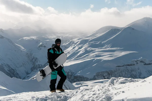 雪人站在雪山上 手里拿着雪板 面对着笼罩在雾蒙蒙的太阳光中的山峰 — 图库照片