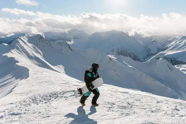 Snowboarder walks on a snowy mountain along a mountain ridge, with a snowboard in his hands against the background of mountain peaks illuminated by the suns rays