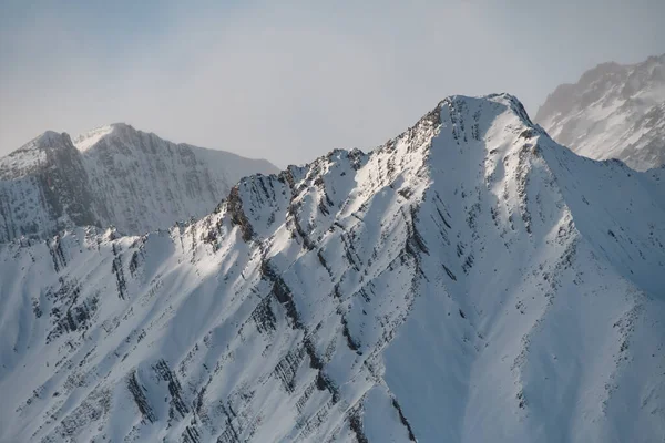 雪で覆われた山頂のクローズアップ写真ですが 岩の黒いアウトクロップは部分的に見え 他のピークは後ろに見えます — ストック写真