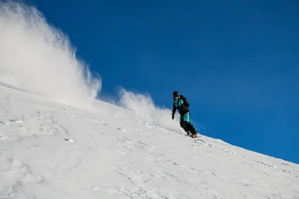 彼のスノーボード上の黒と青のスキースーツの片腕の男性スノーボーダーは 滑らかな雪の斜面上のルートを克服し 彼の目覚めに粉雪が上昇する — ストック写真