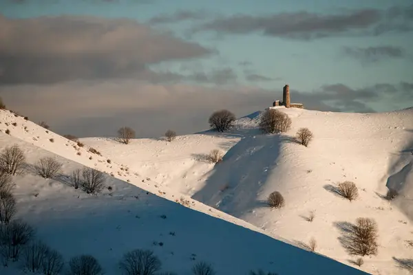 雪地的斜坡 树上长满了树 一座孤零零的古建筑 顶上有一根石柱 阳光照亮 天空阴云密布 — 图库照片