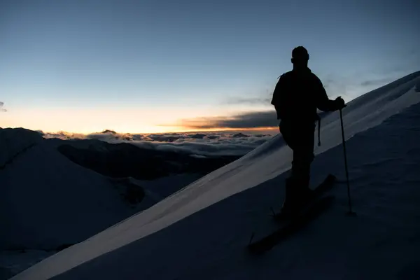 Silhouette of a skier climbing a snowy slope and admiring the peaks of the mountains drowned in white clouds on the background of the sunset