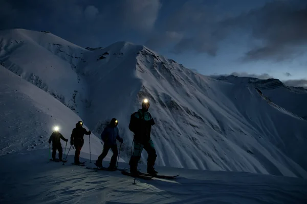 黄昏时分 四个全副武装的滑雪者在雪地的斜坡上滑行 在山水的衬托下 头灯闪烁着光芒 — 图库照片