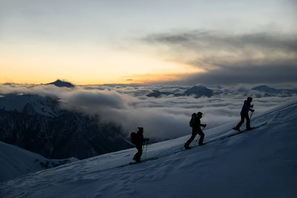 三个滑雪者的轮廓 他们的两根杆子都在山坡上 背靠着落雾覆盖的山峰 — 图库照片