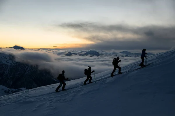 日没後に霧で覆われた山の背景に対して斜面を歩く棒を持つ4人のスキーヤーのシルエット — ストック写真
