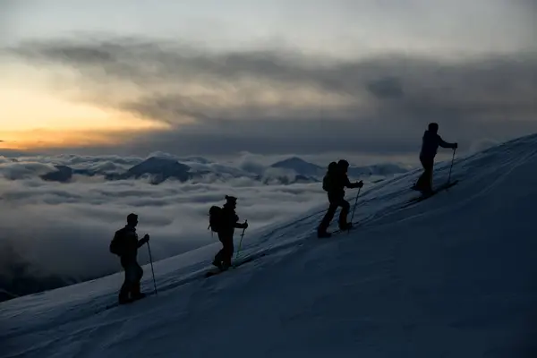 Silhouettes of four skiers with poles climbing to the highest point of a mountain massif against the background of mountains covered with fog after sunset